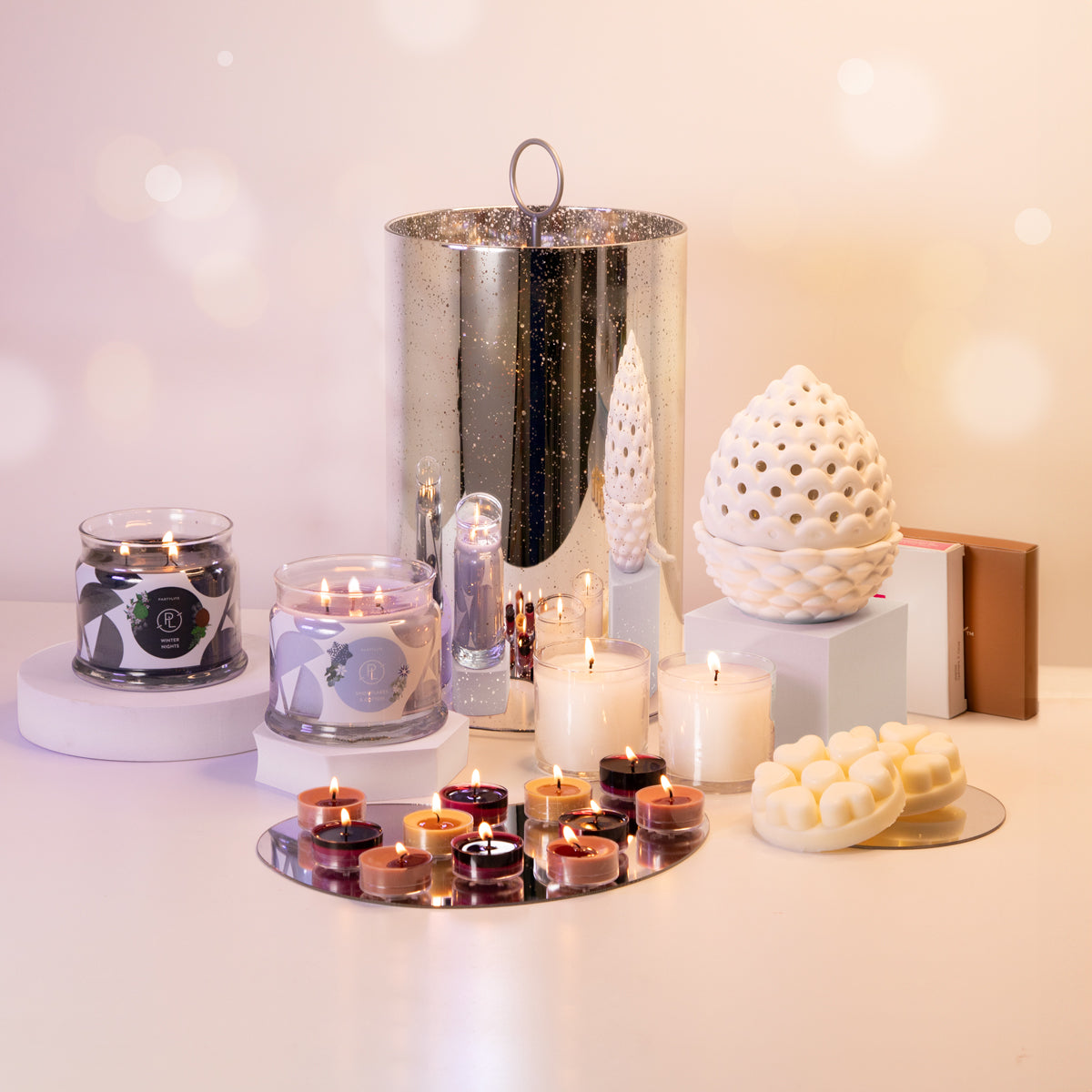 A collection of PartyLite candles and holders displayed with holiday decorations, featuring various tealights, a mercury glass hurricane, and a pinecone scented warmer.