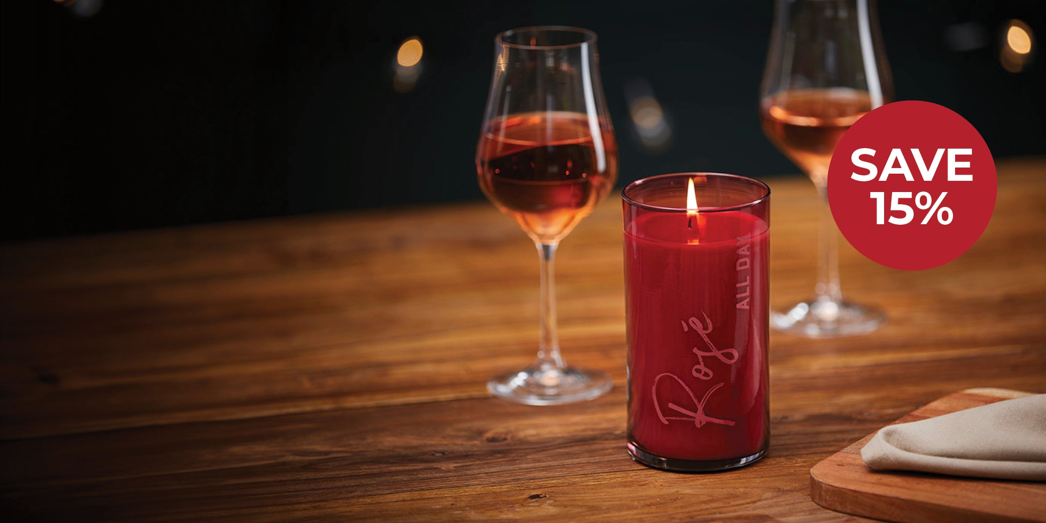 Red candle with 'Rose' branding on a wooden table with wine glasses and a discount label.