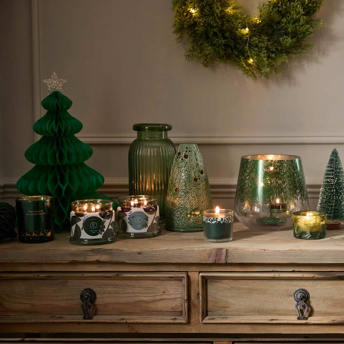 Highland Pine Mini Jar Candle lit with 3-wick jar candles also lit, the Lacy Green Tealight holder and Hurricane with lit tealights, surrounded by Holiday themed items on a wooden cabinet.