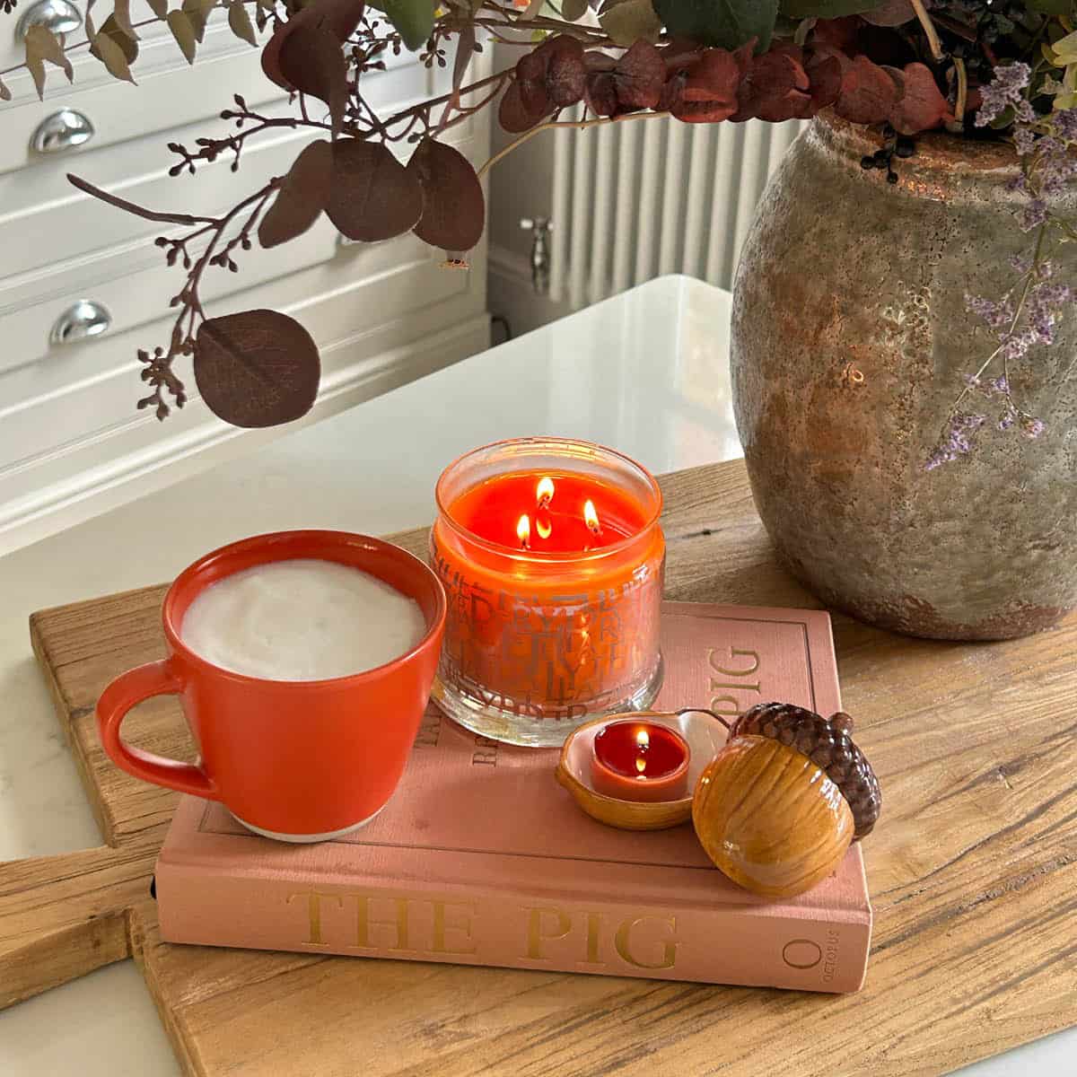 Leaves of Fun 3-Wick Jar Candle lit shown with a mug of hot drink on top of a book with a vase of leaves next to them.