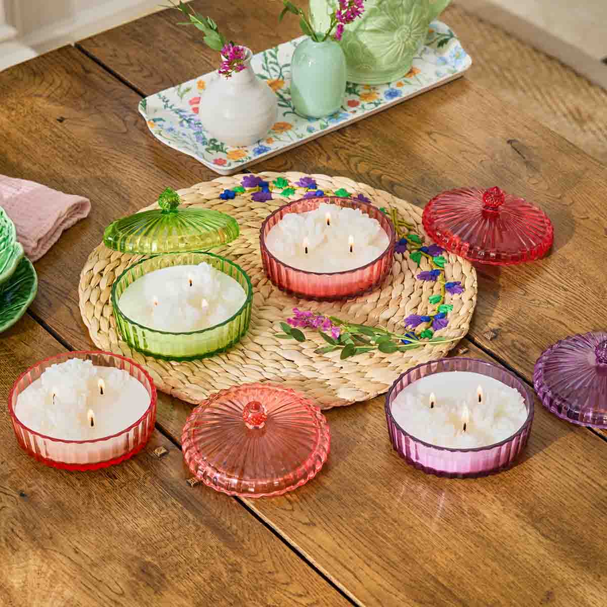 Assortment of Garden Party 3-Wick  colorful Jar Candles on a woven table mat on a wooden table with decorative items in the background.