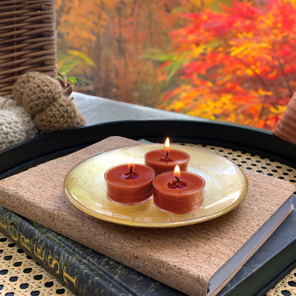 Three Maple Tabac Tealight candles on a gold plate with a blurred autumn scene in the background.