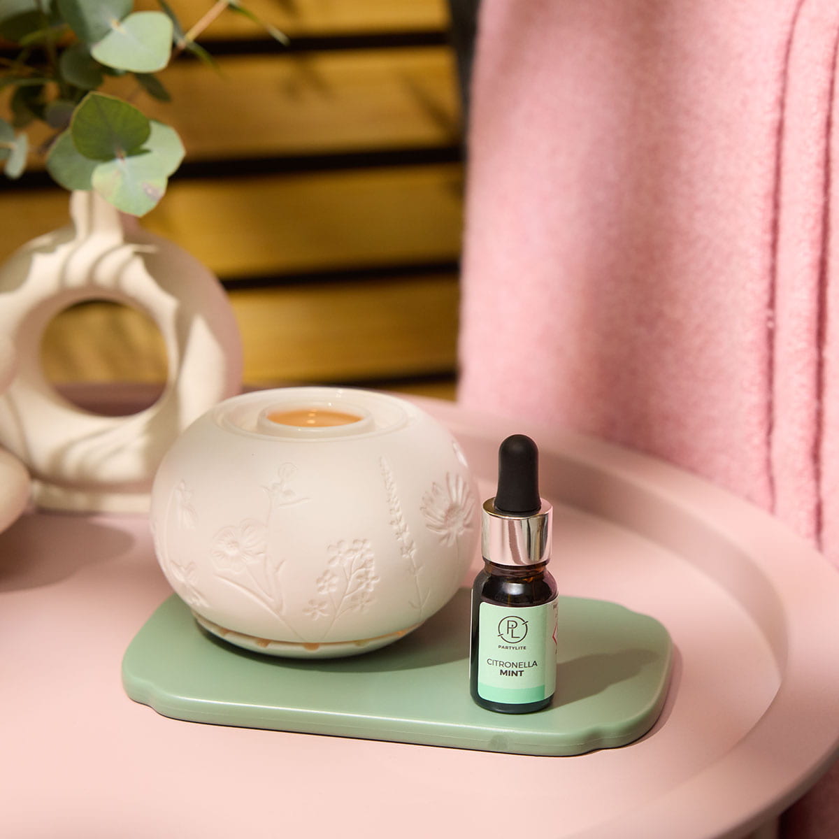 Citronella Mint Fragrance Oil shown with the Illuminescents™ Blooming Fields Fragrance Warmer on a green tray on a pale pink table with a pink blanket in the background and a decorative plant 